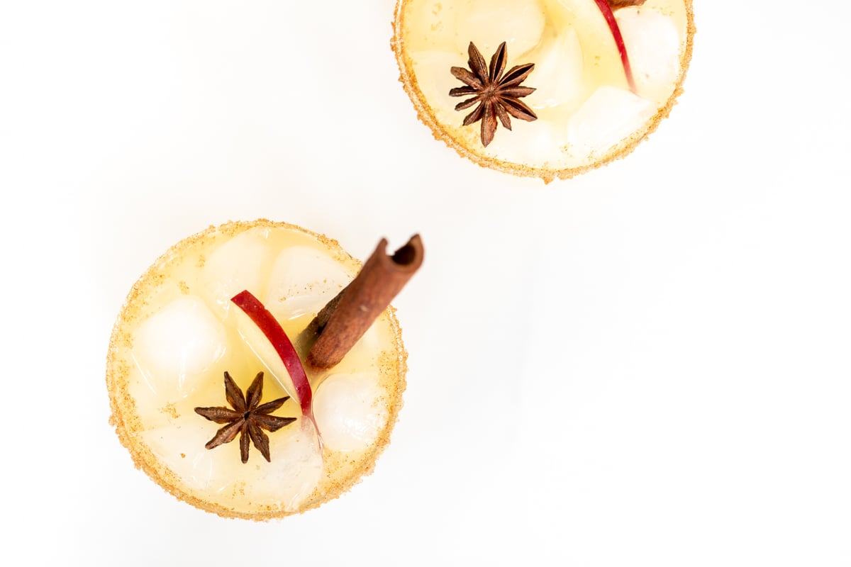 Clear glasses with apple cider margaritas, rimmed in sugar and garnished with a cinnamon stick on a marble surface.