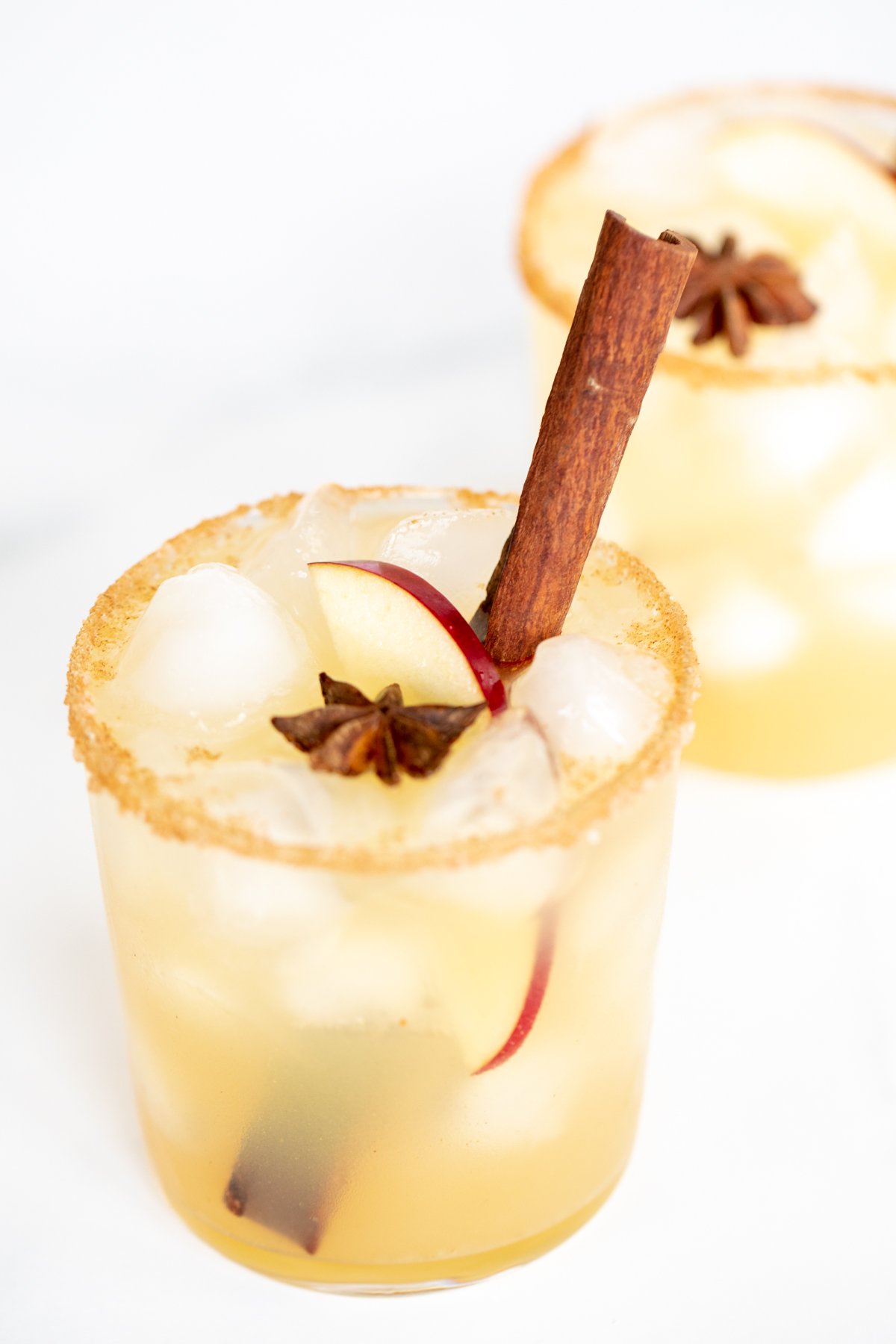 Clear glasses with apple cider margaritas, rimmed in sugar and garnished with a cinnamon stick on a marble surface.