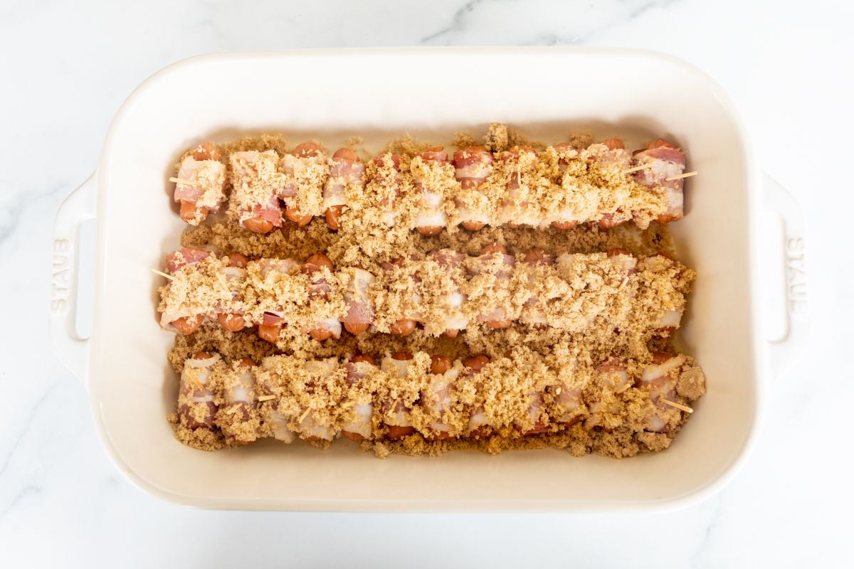 A white baking dish lined with bacon wrapped brown sugar little smokies before baking.