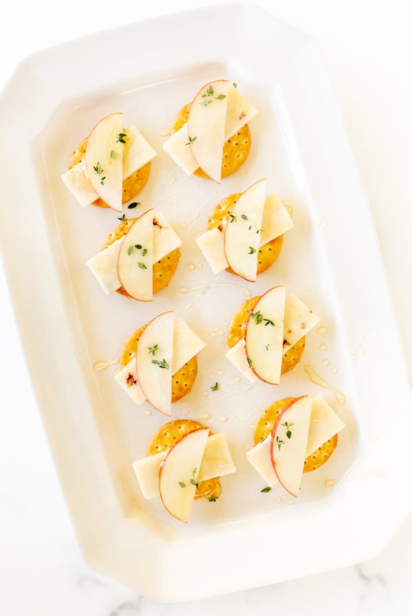 Cheese and crackers on a white platter, with an apple slice and fresh herbs.