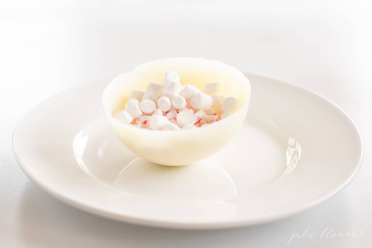 A white chocolate hot cocoa bomb on a white plate, split in half to show marshmallows and mix.