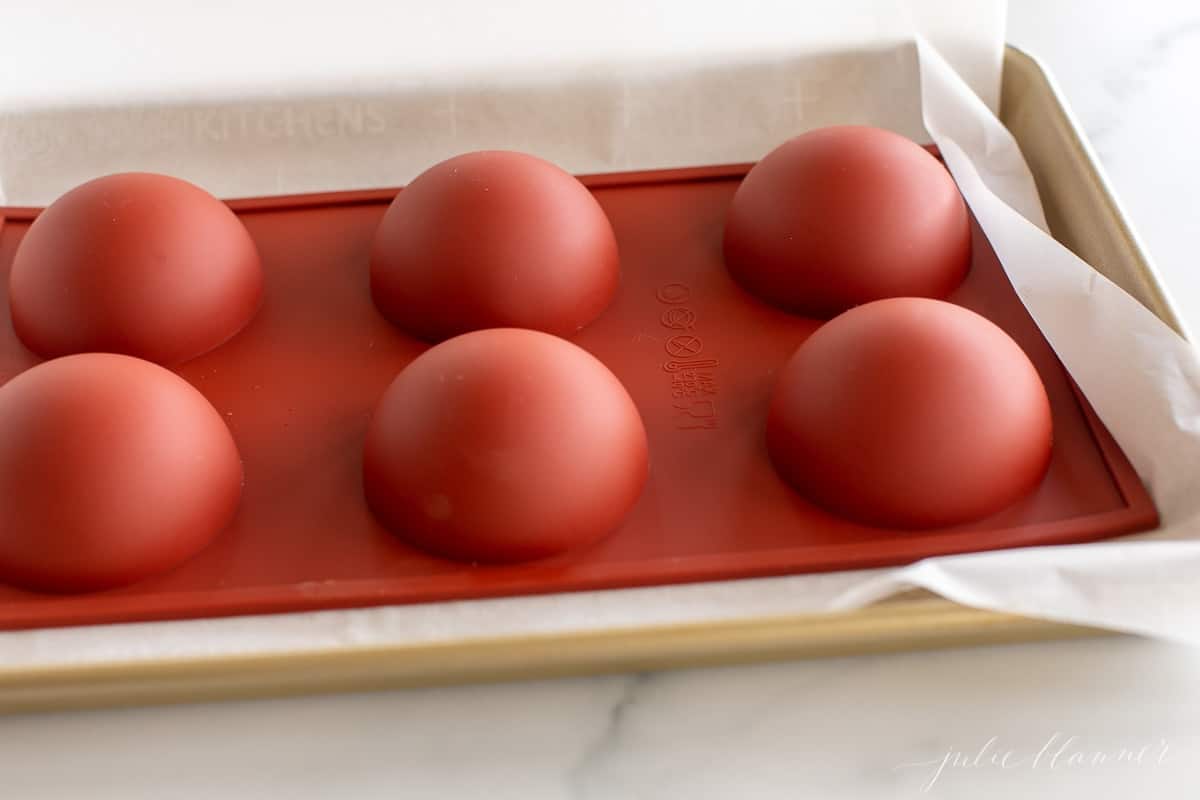 A red silicone sphere mold filled with chocolate for Mexican hot chocolate bombs, inverted on a gold baking sheet.