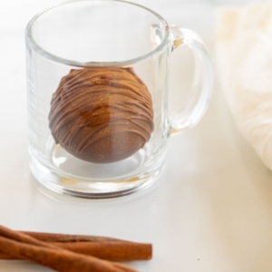 A Mexican hot chocolate bomb in a clear glass mug, with cinnamon sticks in the foreground.