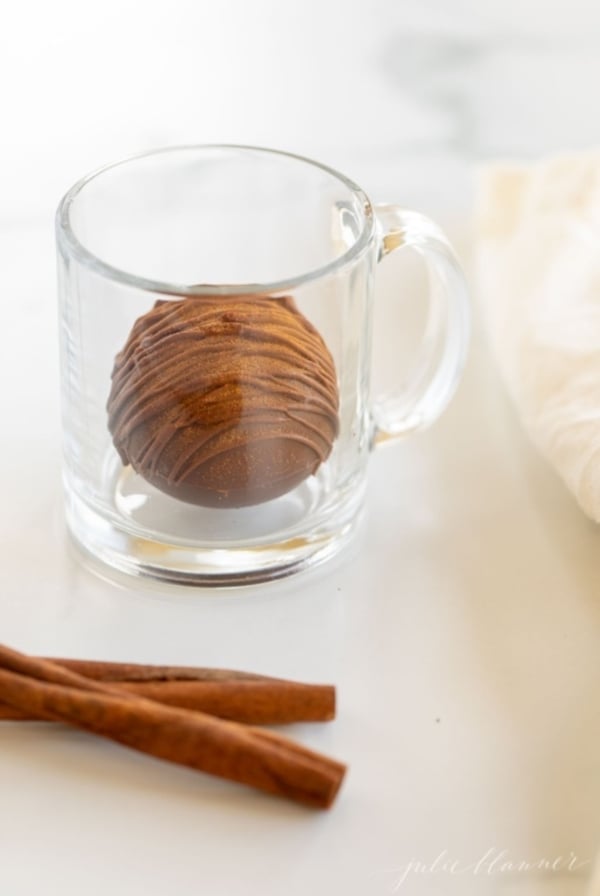 A Mexican hot chocolate bomb in a clear glass mug, with cinnamon sticks in the foreground.