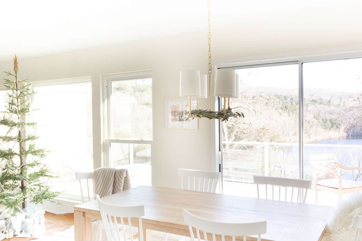 A soft wood kitchen table with white chairs and a Scandinavian tree in the background, lake view through the windows.