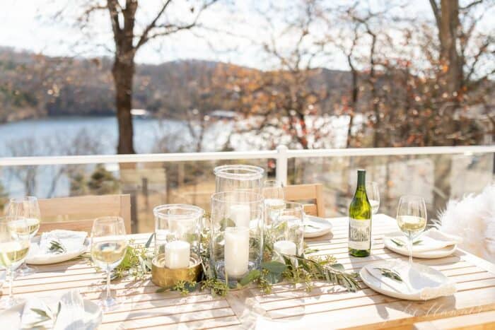 An outdoor table set for a Christmas dinner with clear glass hurricanes and greenery.