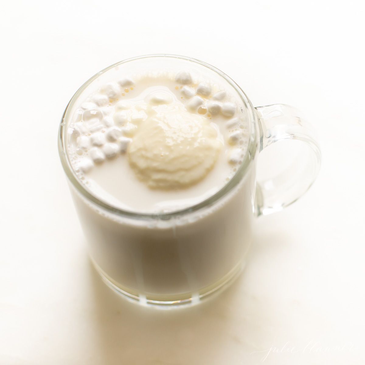 A clear mug of white hot chocolate on a marble countertop.