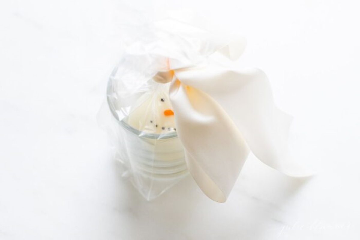 A clear glass mug with a snowman hot chocolate mug inside, tied with a white satin bow.