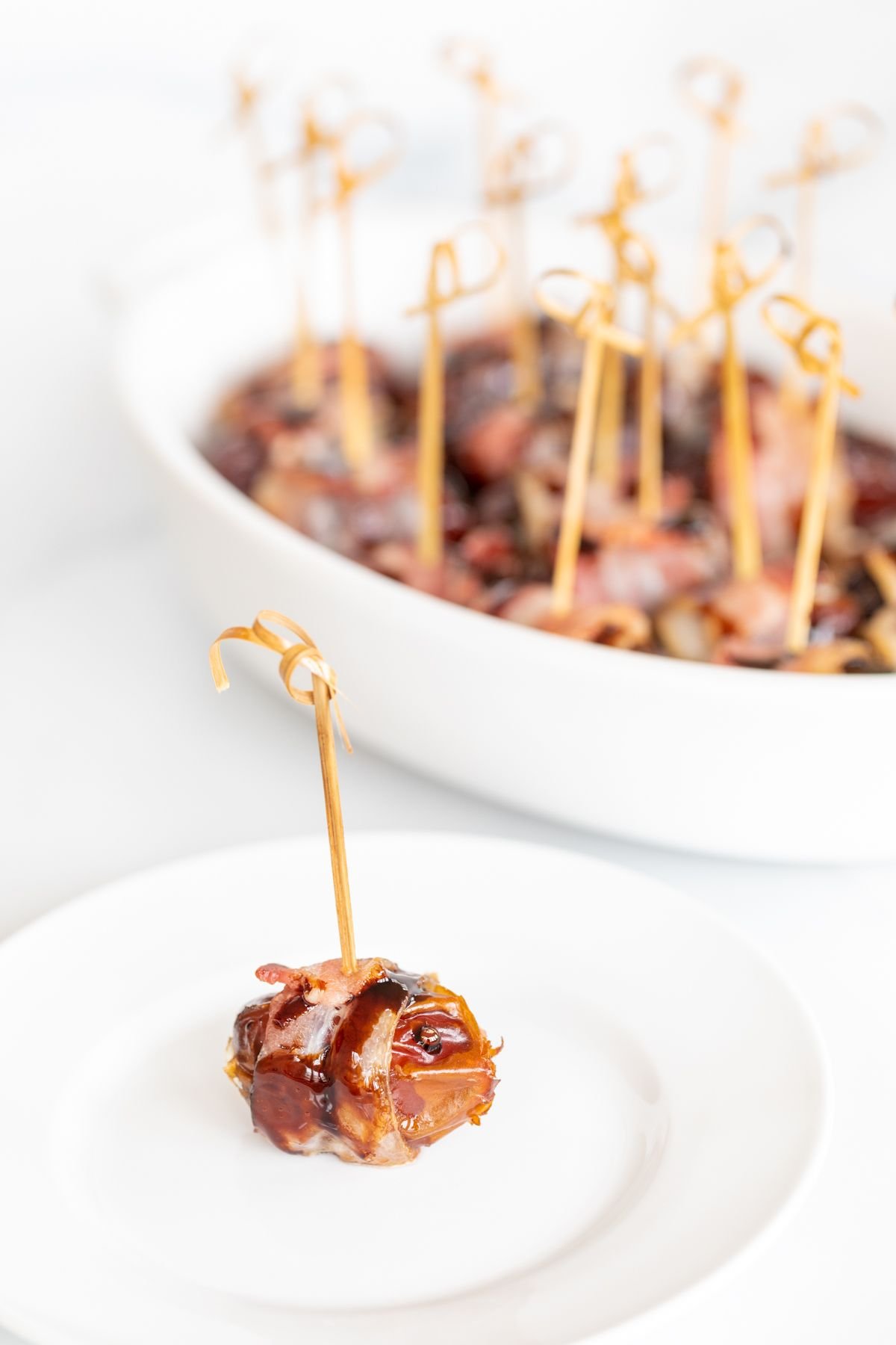 A single stuffed date wrapped in bacon on a white plate, with a dish full of them in the background.