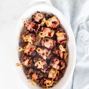 A white oval baking dish with bacon wrapped dates, each has a toothpick for serving.