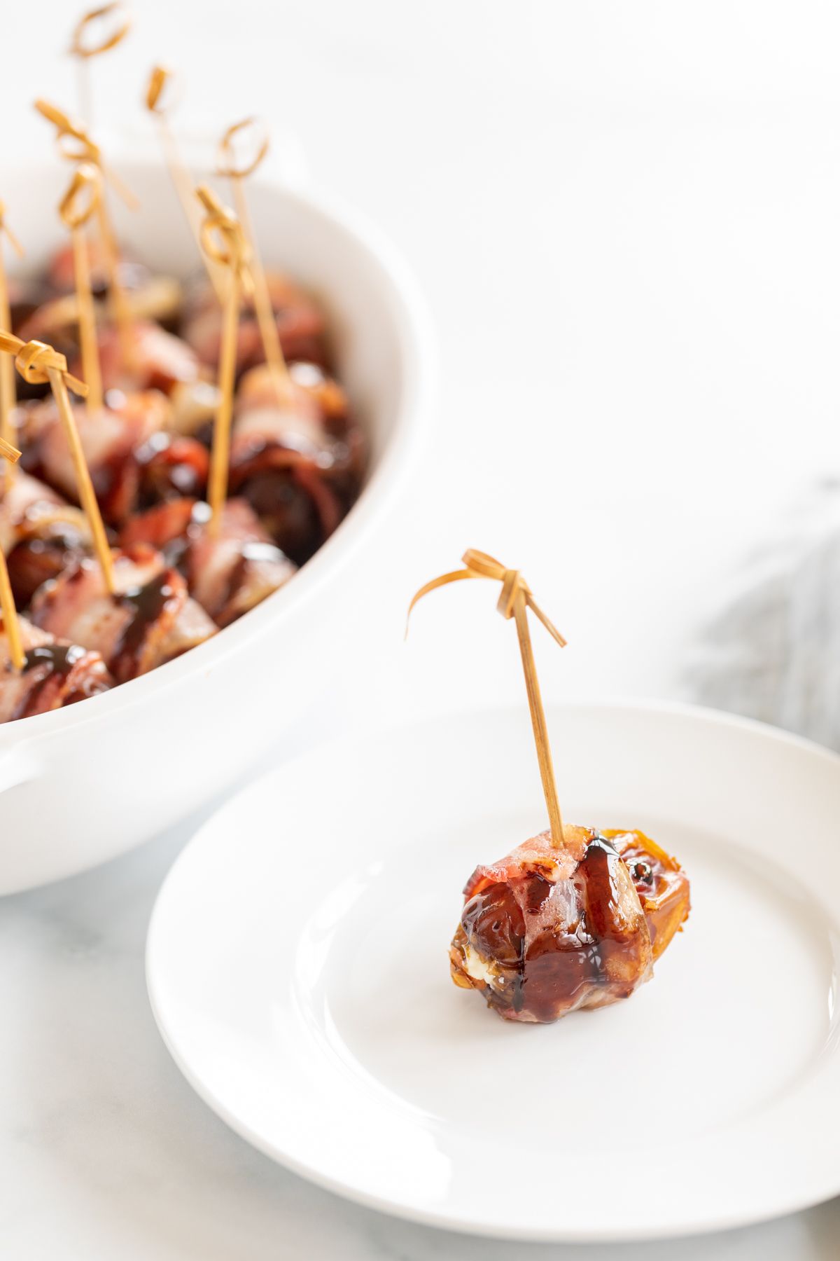 A single stuffed date wrapped in bacon on a white plate, with a dish full of them in the background. 