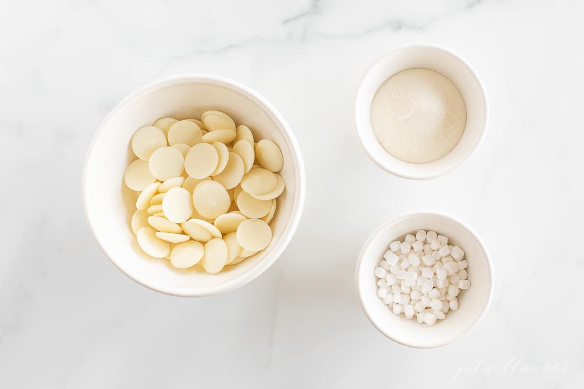 A marble surface with small white bowls filled with ingredients for white chocolate hot chocolate bombs.