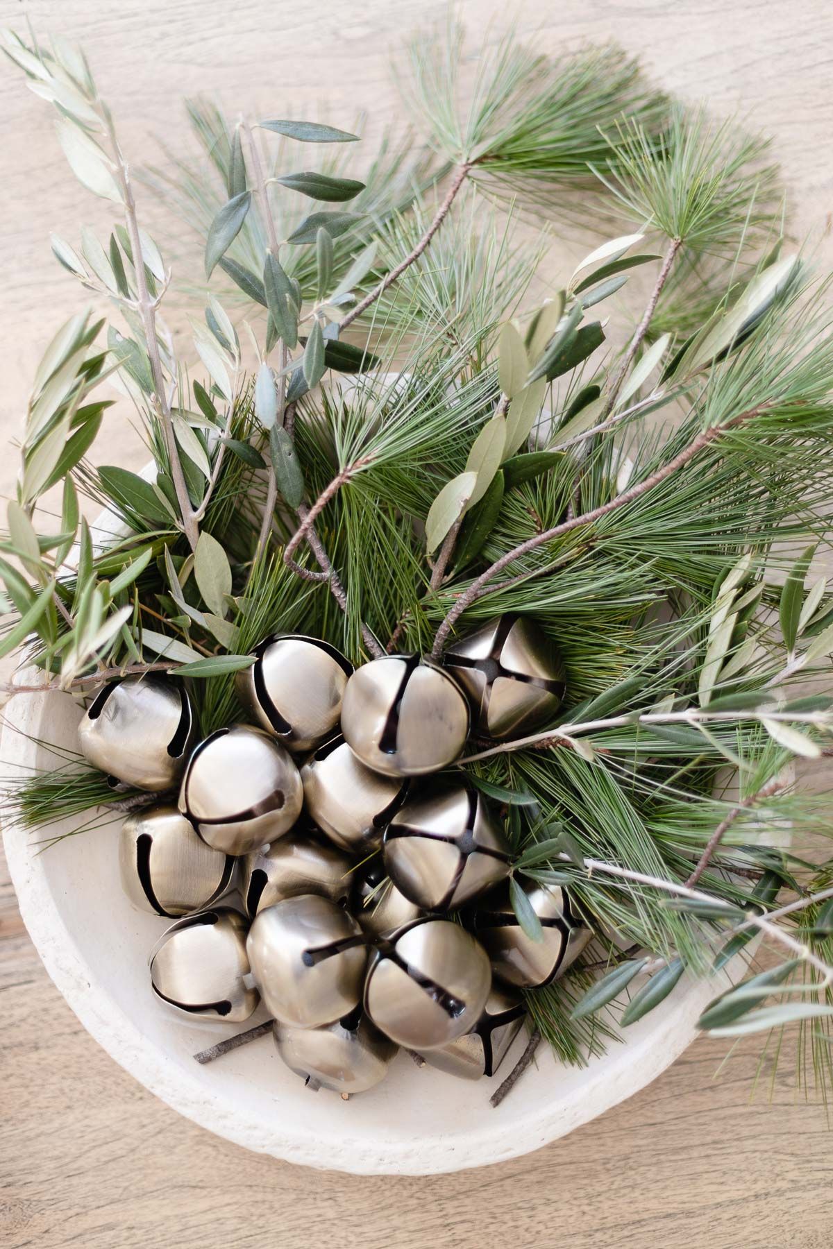 A bowl of silver jingle bells and fresh greenery on a wood table