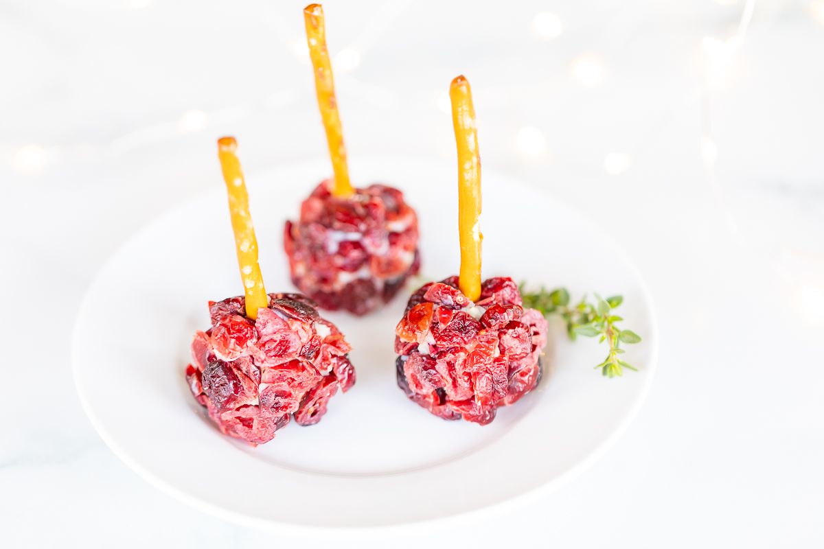 Mini cheese ball bites covered in dried cranberries, with a pretzel stick in the center, resting on a white platter.