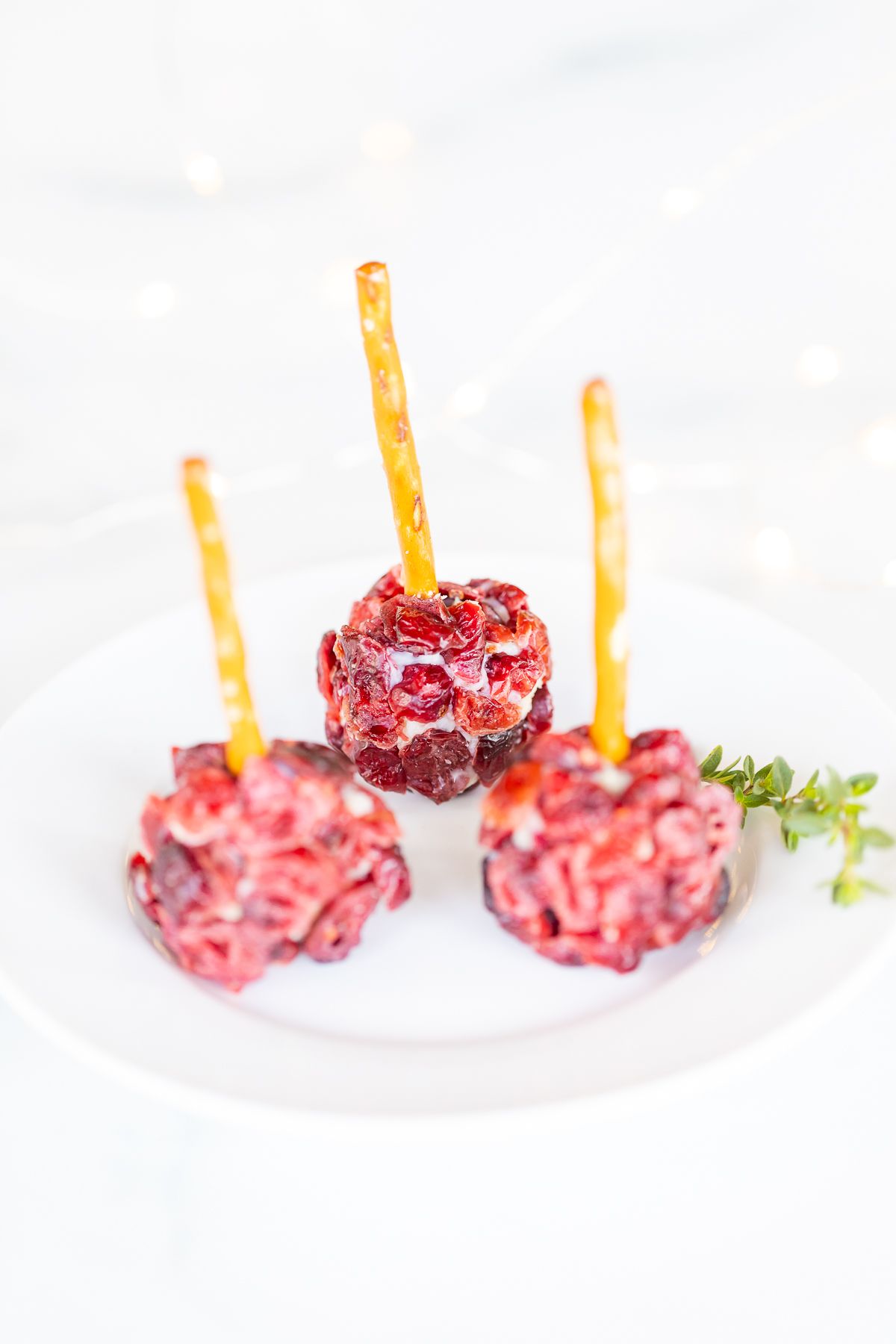 Mini cheese ball bites covered in dried cranberries, with a pretzel stick in the center, resting on a white platter.