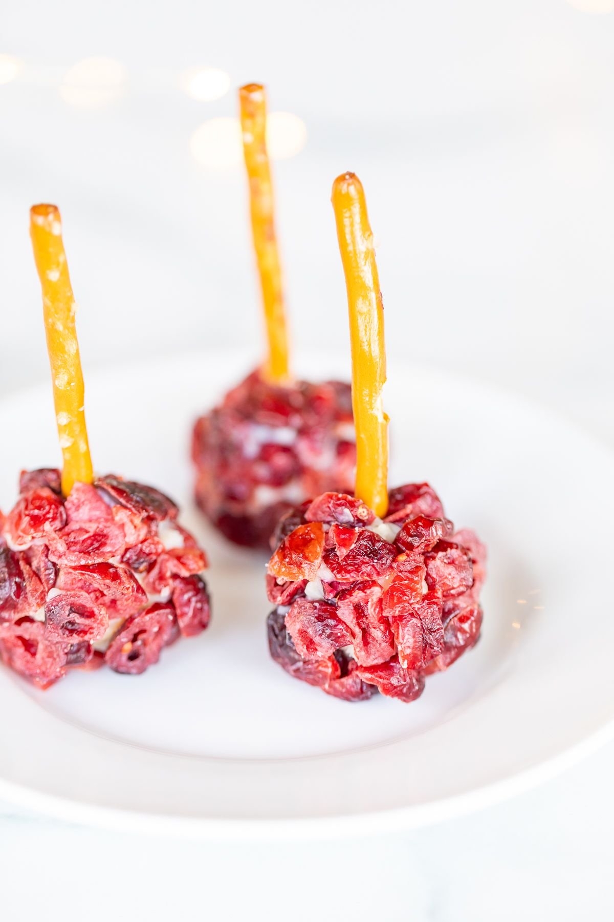 Mini cheese ball bites covered in dried cranberries, with a pretzel stick in the center, resting on a white platter.