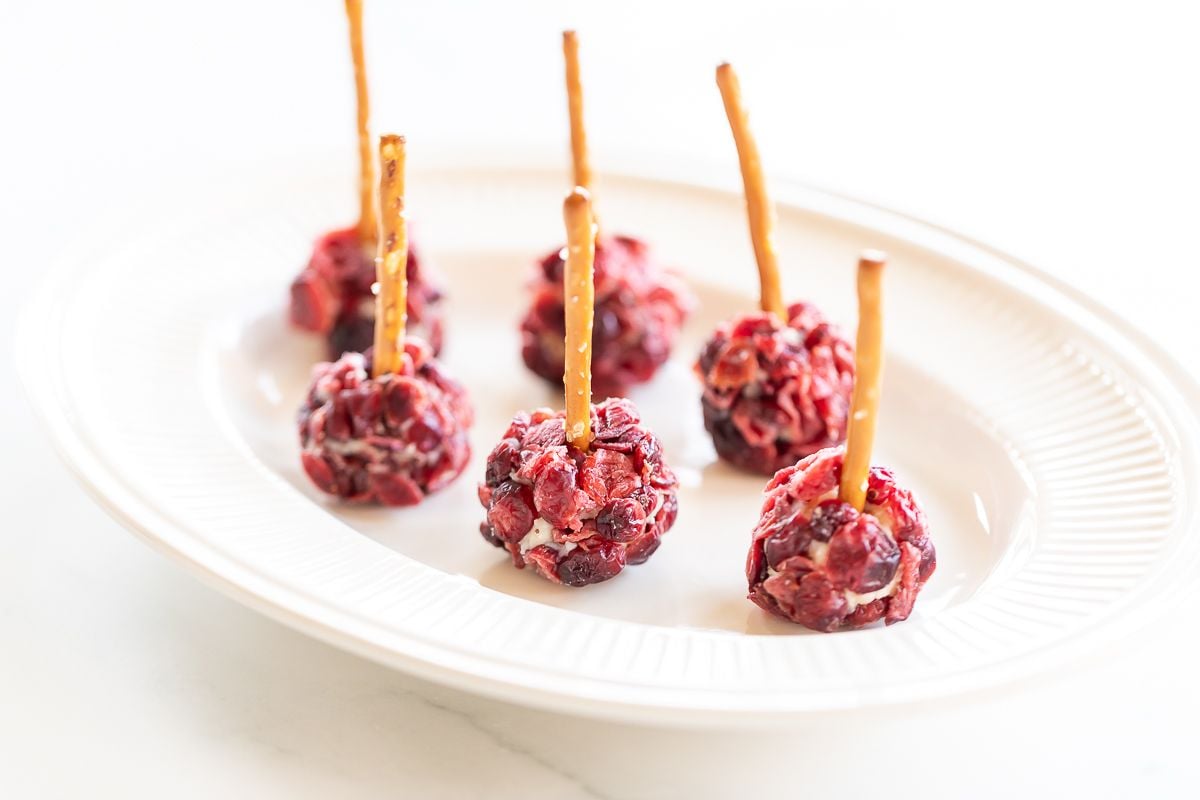 Mini cheese ball bites covered in dried cranberries, with a pretzel stick in the center, resting on a white platter.