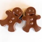 A pair of dark chocolate hot chocolate bombs shaped as gingerbread men on a white marble surface.