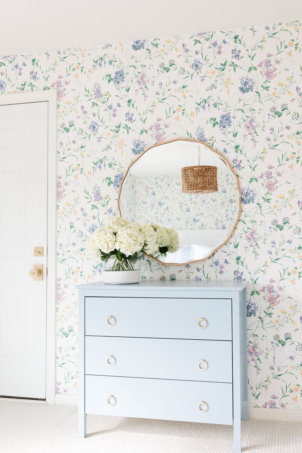 A guest bedroom with flowered wallpaper, a wavy mirror and flowers on top of a blue dresser.