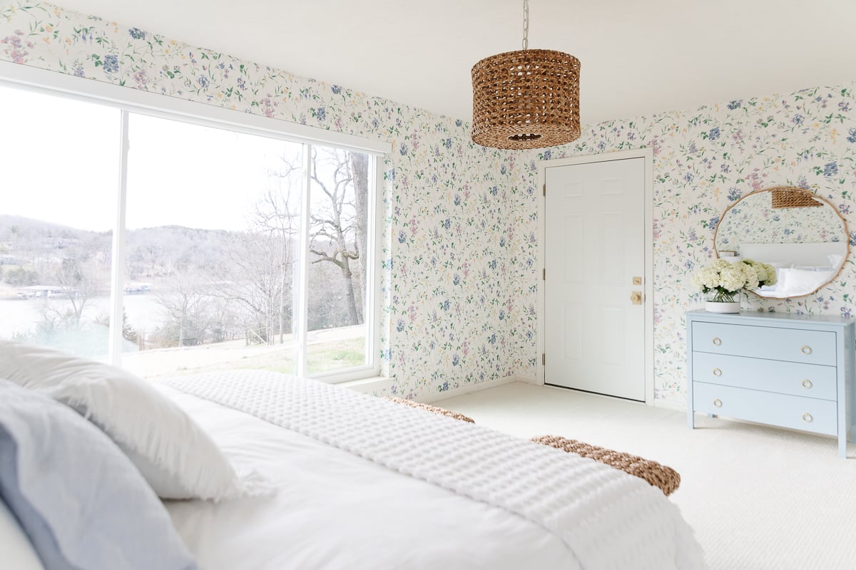 A wallpapered guest bedroom with white bedding and rattan stools.