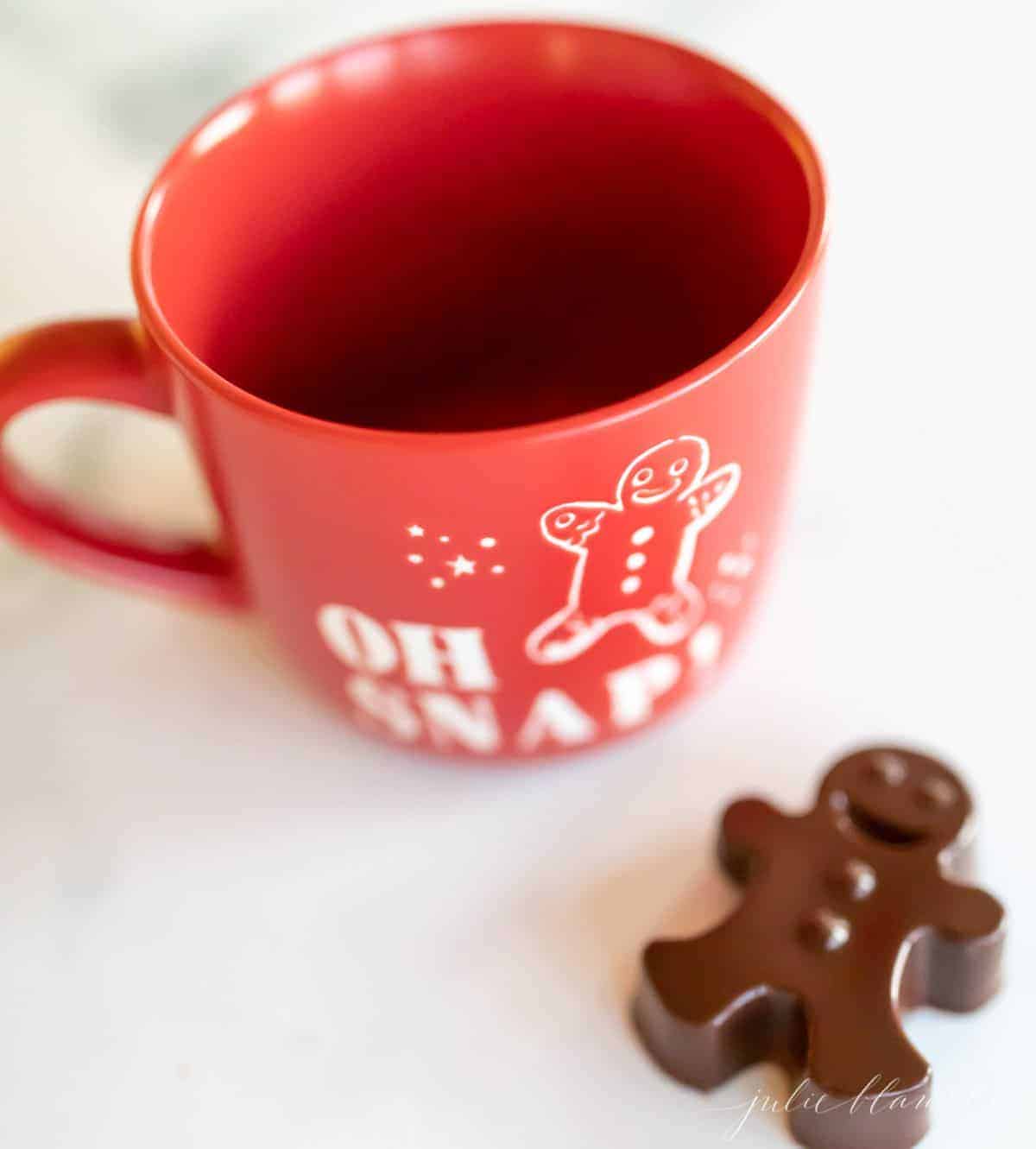 A red mug for hot chocolate, with a dark hot chocolate bomb shaped as a gingerbread man to the side.