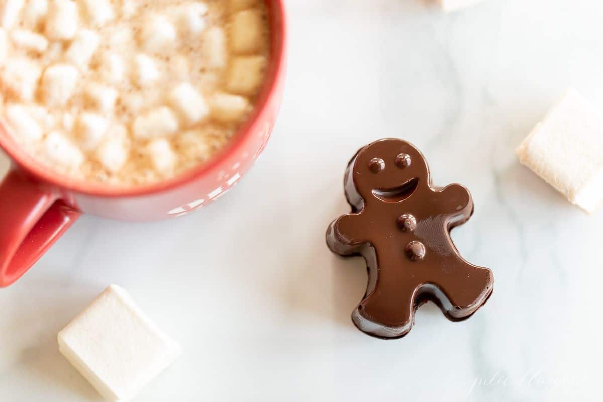 A red mug full of dark hot chocolate with marshmallows, with a dark chocolate gingerbread man to the side.