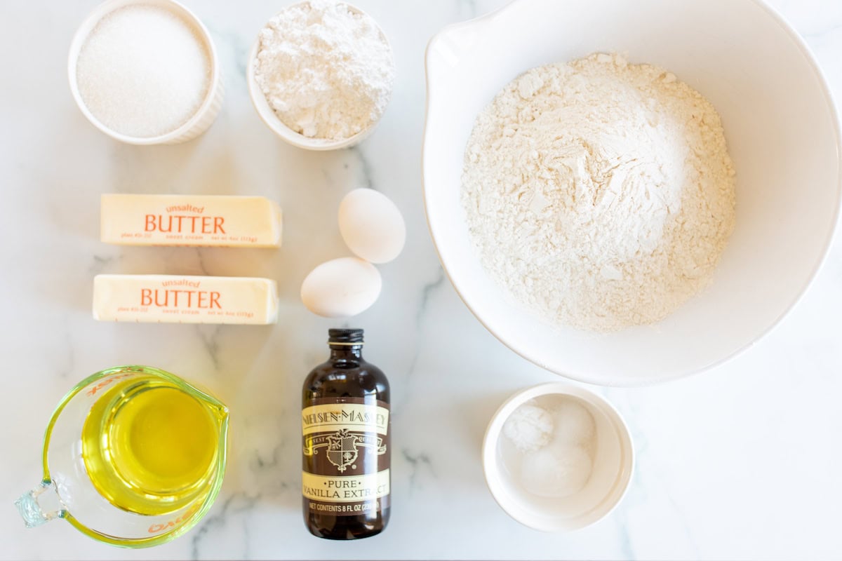 A collection of baking ingredients for Amish sugar cookies, including two sticks of butter, two eggs, flour, sugar, baking soda, baking powder, vanilla extract, and oil displayed on a white surface.