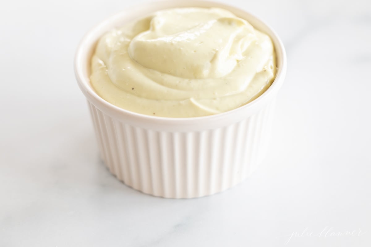 A white ramekin full of homemade avocado mayo, on a marble countertop.