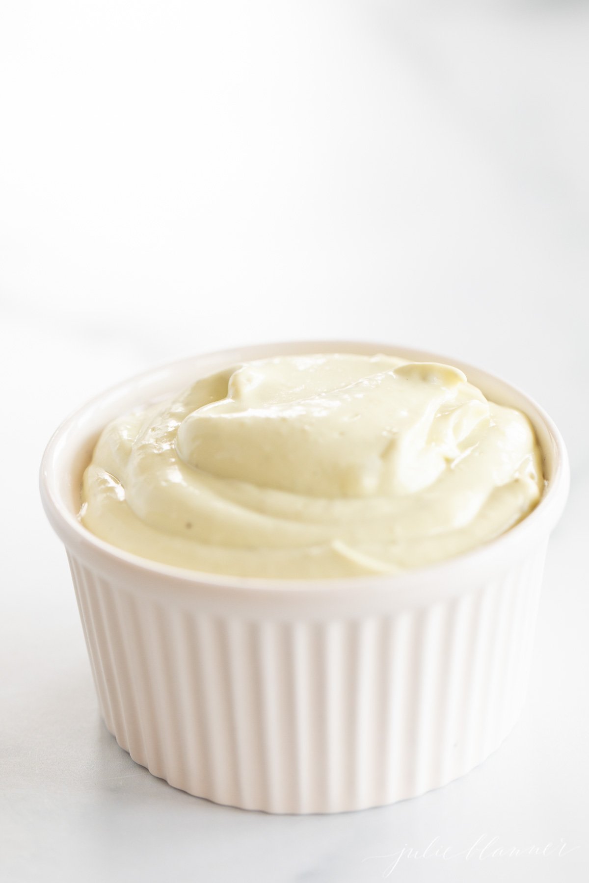 A white ramekin full of homemade avocado mayo, on a marble countertop.