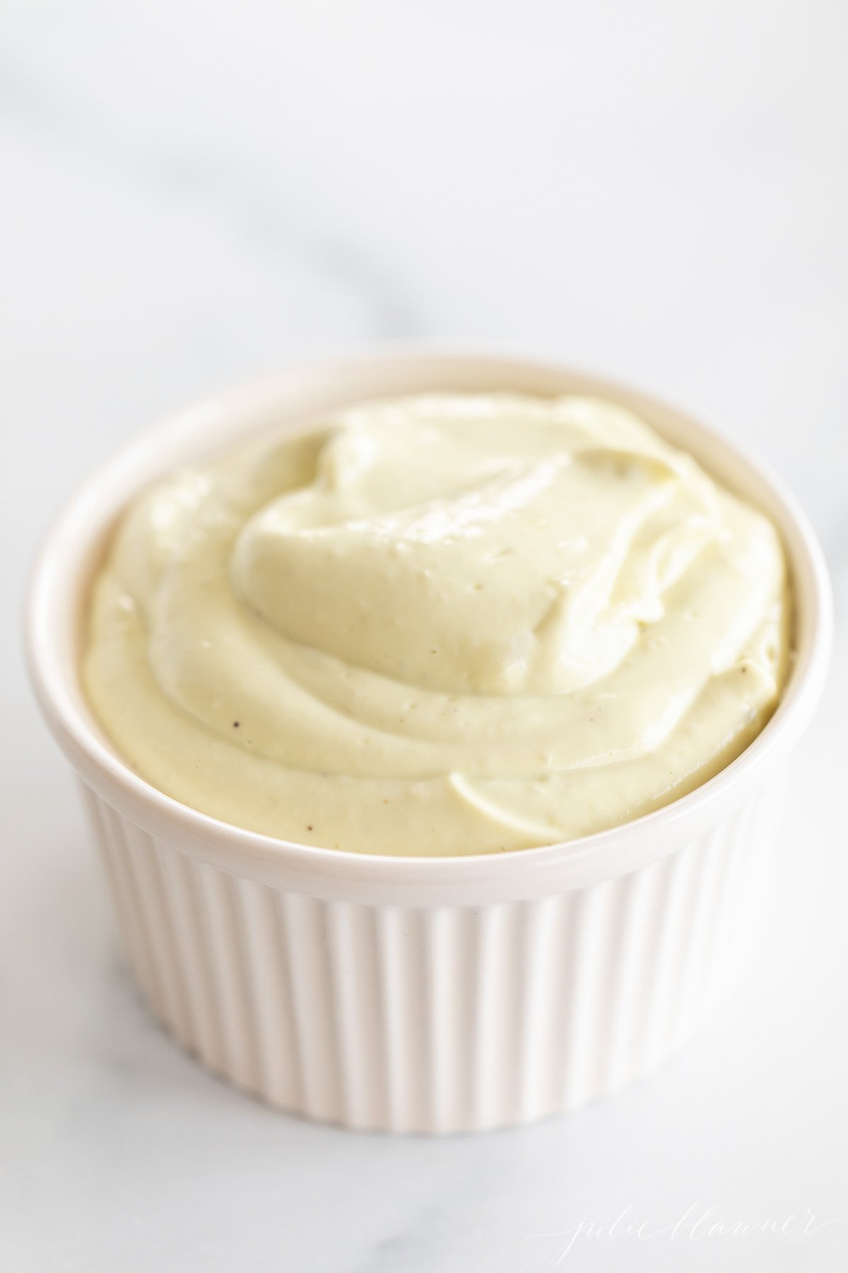 A white ramekin full of homemade avocado mayo, on a marble countertop.