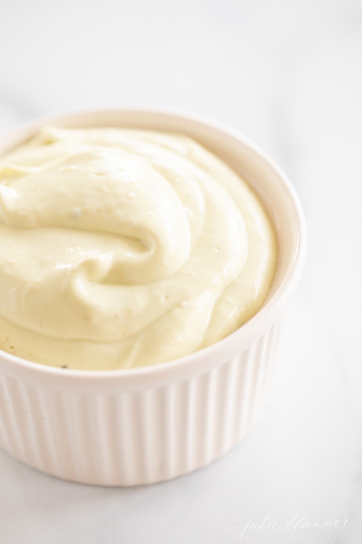 A white ramekin full of homemade avocado mayo, on a marble countertop.