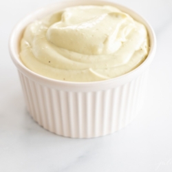 A white ramekin full of homemade avocado mayo, on a marble countertop.