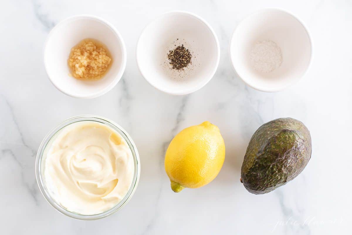 Small bowls of ingredients on a marble surface for an avocado mayo recipe.