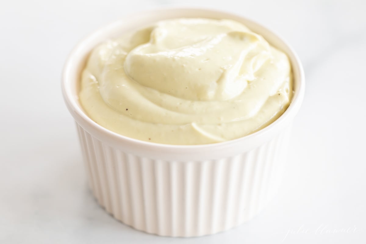 A white ramekin full of homemade avocado mayo, on a marble countertop. 