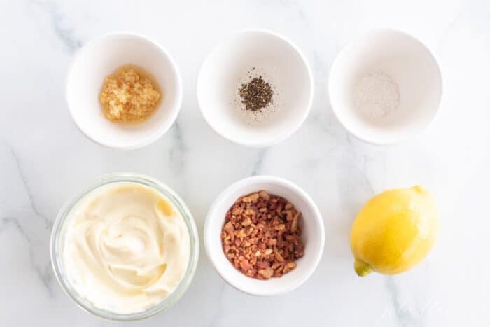 Small white bowls of ingredients for a bacon aioli recipe laid out on a marble surface.