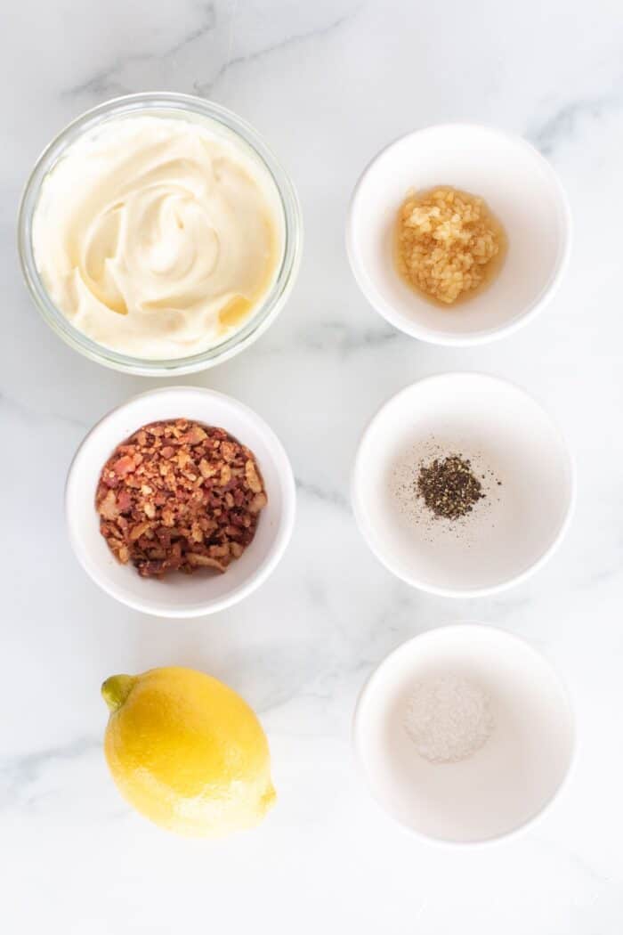 Small white bowls of ingredients for a bacon aioli recipe laid out on a marble surface.