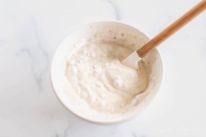 A white mixing bowl full of bacon mayo on a marble surface, spatula in the bowl.