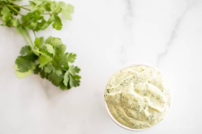 A white bowl full of cilantro aioli with a piece of fresh cilantro on a marble surface.