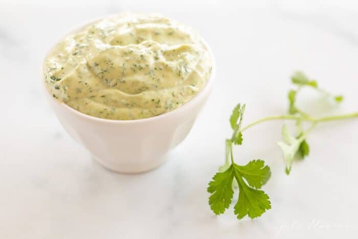 A white bowl full of cilantro lime aioli with a piece of fresh cilantro on a marble surface.