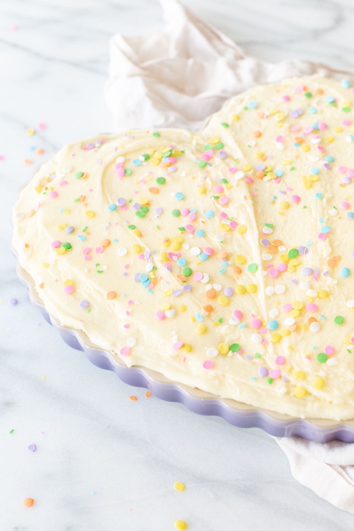 A frosted cookie cake topped with sprinkles, in a heart shaped pan, on a marble countertop.