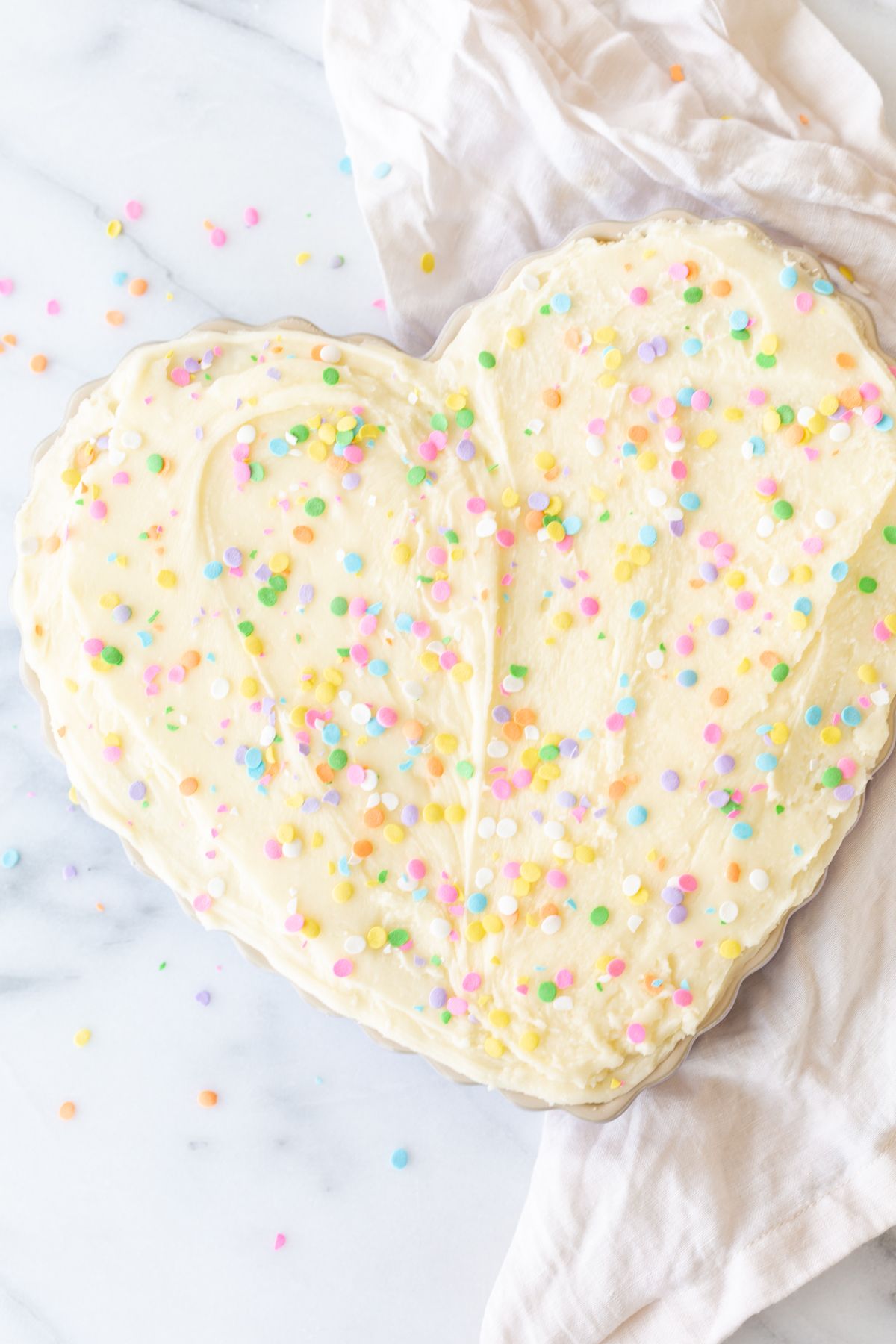 A frosted cookie cake topped with sprinkles, in a heart shaped pan, on a marble countertop.