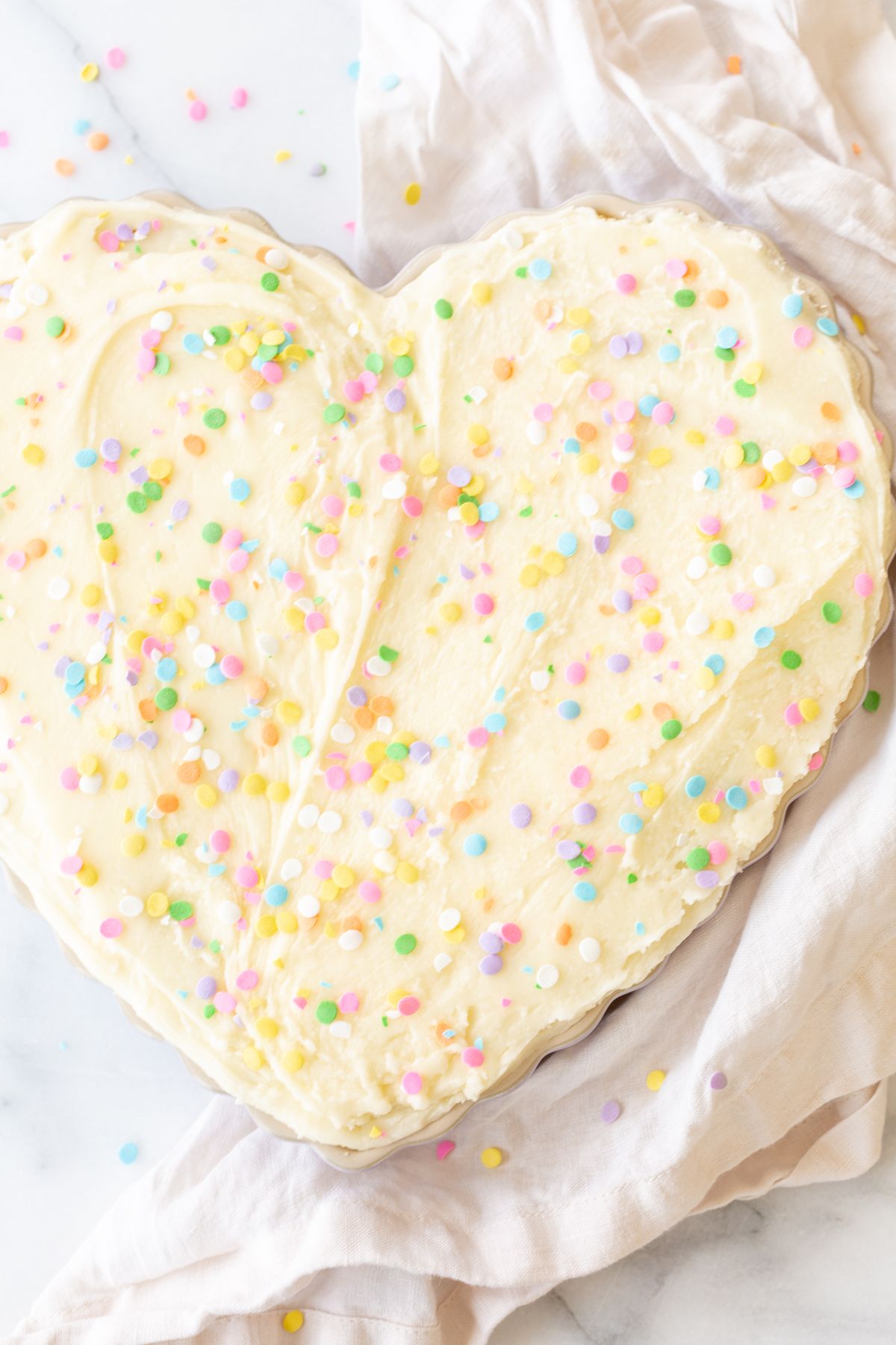 A frosted cookie cake topped with sprinkles, in a heart shaped pan, on a marble countertop.