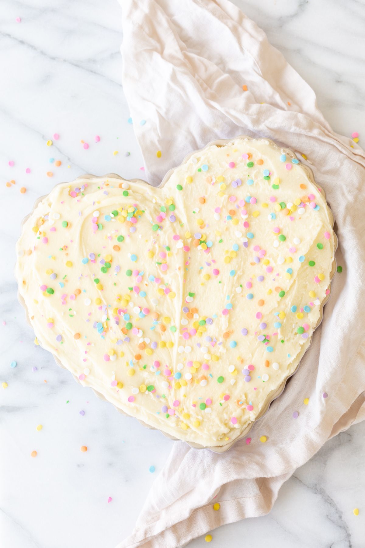 A frosted cookie cake topped with sprinkles, in a heart shaped pan, on a marble countertop.