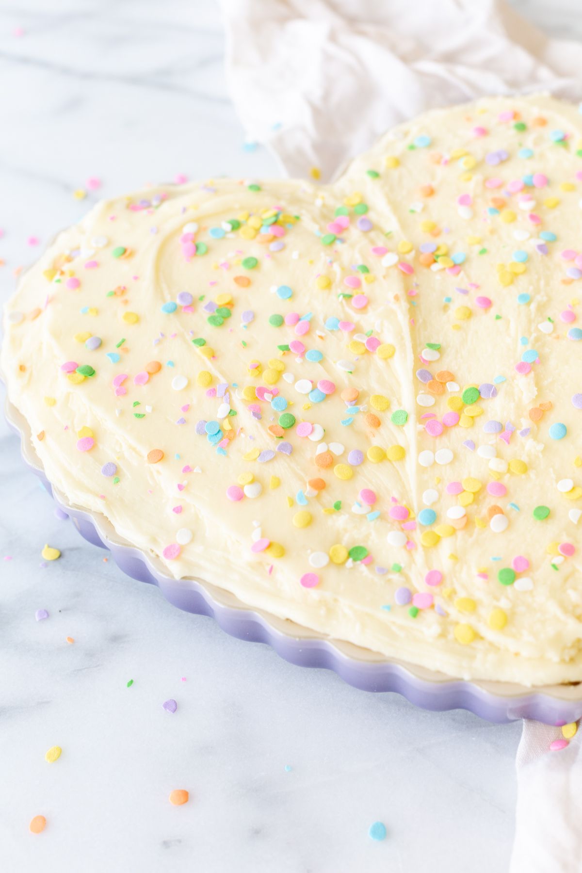 A frosted cookie cake topped with sprinkles, in a heart shaped pan, on a marble countertop.
