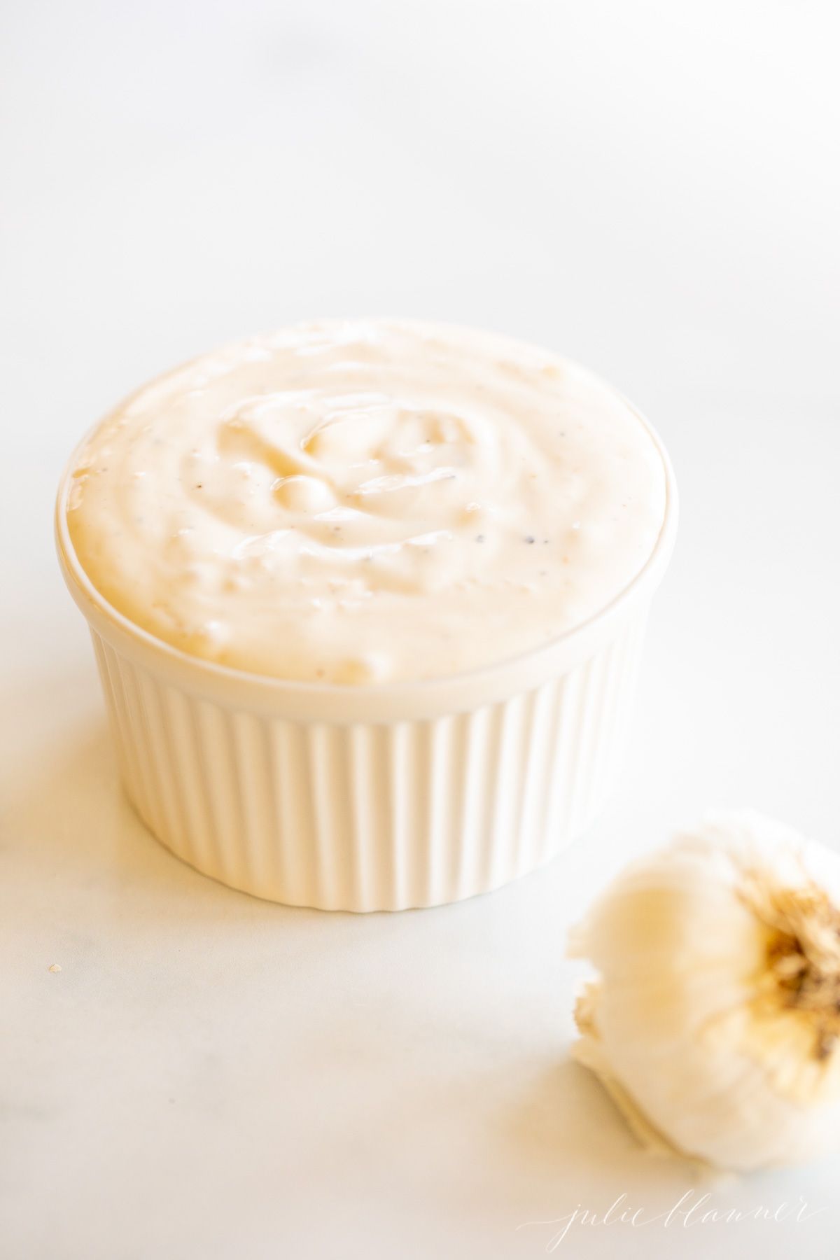 A small white bowl of garlic aioli with a garlic bulb to the side.