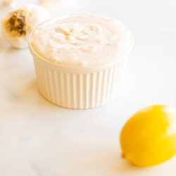 A white ramekin full of garlic aioli with a garlic bulb and a lemon in the foreground.