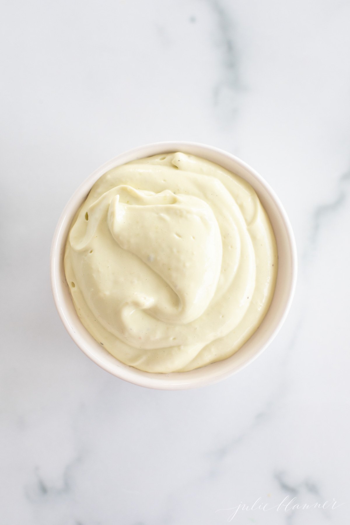 A white ramekin full of homemade avocado mayo, on a marble countertop.