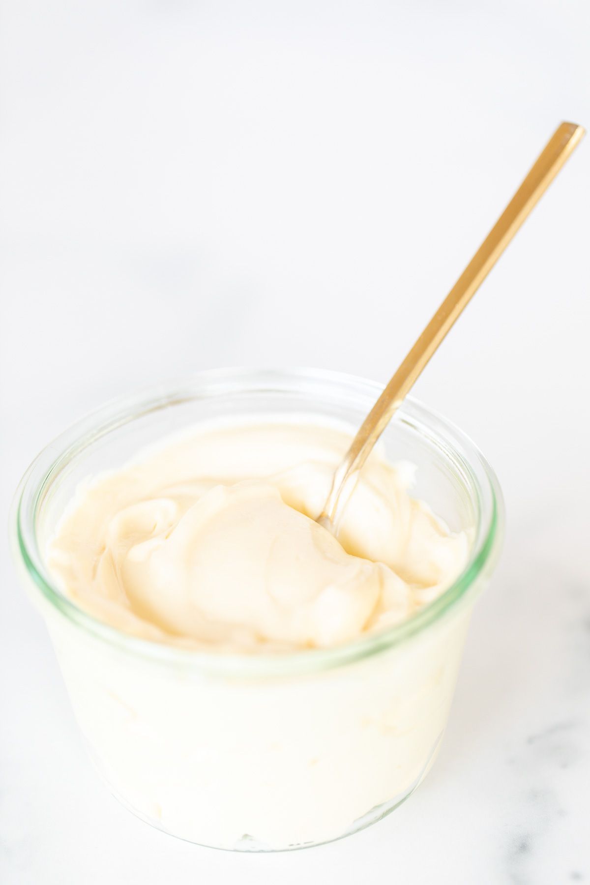 A small glass container of homemade mayonnaise with a gold spoon, resting on a marble countertop.