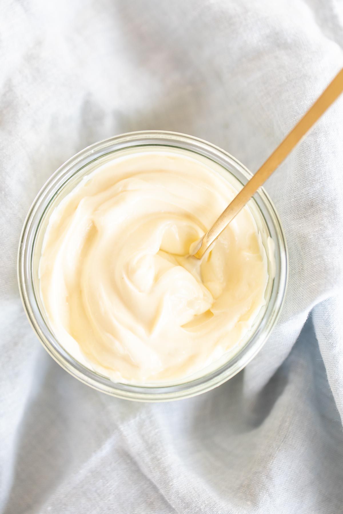 A small glass container of homemade mayonnaise with a gold spoon, resting on a marble countertop.