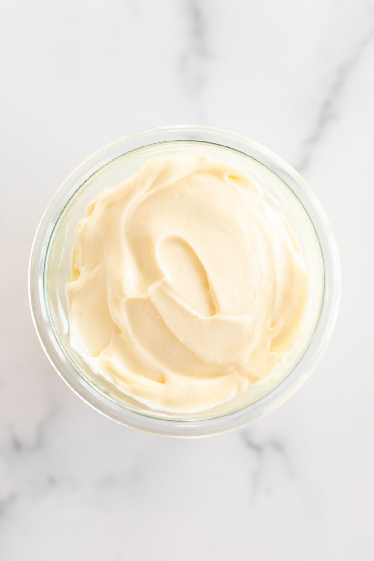 A small glass jar of mayonnaise for a tutorial on how to make mayo, it's resting on a marble countertop.